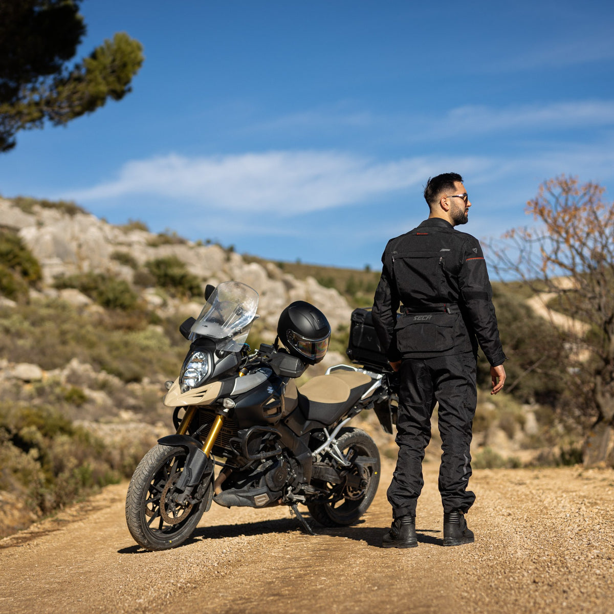 Man klädd i Seca Textil Mc-Jacka Discovery Svart bakifrån bredvid en motorcykel, visar jackans passform och funktionalitet.